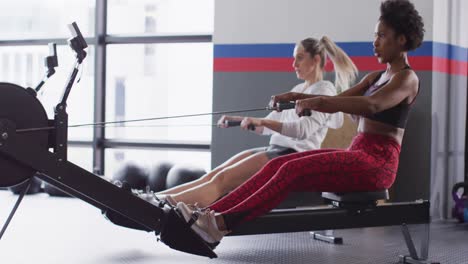 Video-of-two-diverse,-confident-women-on-rowing-machines-working-out-at-a-gym