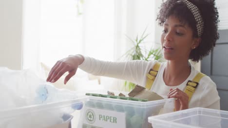 Video-of-happy-biracial-woman-sorting-recycling-and-smiling-at-home,-with-copy-space