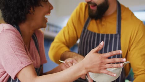 Video-of-laughing-diverse-couple-in-aprons-talking-and-baking-together-in-kitchen-at-home
