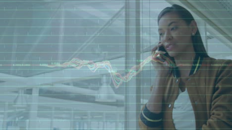 Animation-of-multicolored-graphs,-numbers,-biracial-woman-talking-on-cellphone-in-corridor-of-office