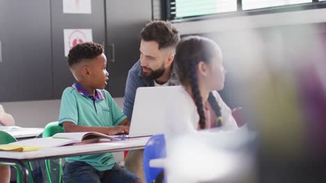 Diverse-male-teacher-and-happy-schoolchildren-using-laptop-in-school-classroom