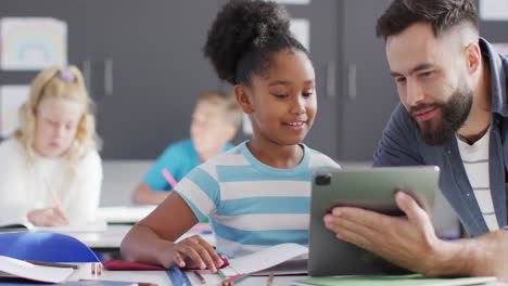 Diverse-male-teacher-and-happy-schoolchildren-using-tablet-in-school-classroom