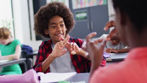 Diverse-female-teacher-and-happy-schoolchildren-at-desks-learning-sign-language-in-school-classroom