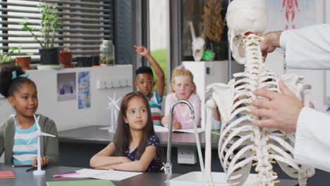 Diverse-male-teacher-and-happy-schoolchildren-answering-questions-about-skeleton-in-biology-class