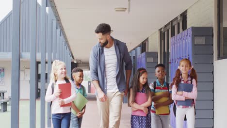 Diverse-male-teacher-and-happy-schoolchildren-walking-at-school