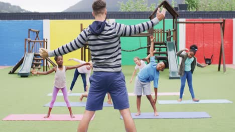 Diverse-male-teacher-and-happy-schoolchildren-exercising-on-mats-at-school-playground