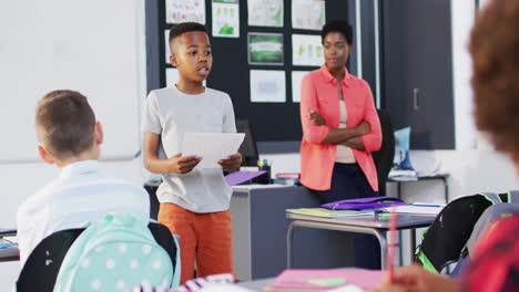 Diverse-female-teacher-and-happy-schoolchildren-at-desks-reciting-in-school-classroom
