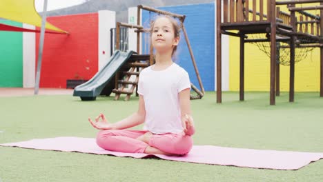 Portrait-of-happy-caucasian-schoolgirl-meditating-on-mat-at-school-playground