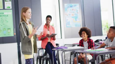 Diverse-female-teacher-and-happy-schoolchildren-at-desks-reciting-in-school-classroom