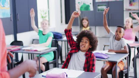 Diverse-female-teacher-and-happy-schoolchildren-at-desks-raising-hands-in-school-classroom
