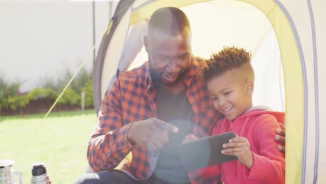 Glücklicher-Afroamerikanischer-Vater-Und-Sein-Sohn-Sitzen-Im-Zelt-Und-Benutzen-Tablet-Im-Garten