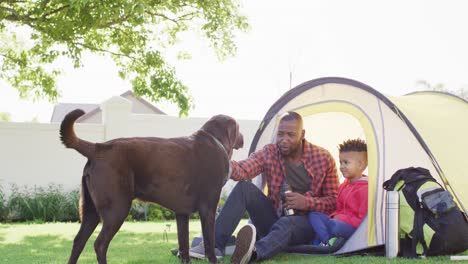 Happy-african-american-father-and-his-son-with-pet-dog-sitting-in-tent-in-garden