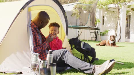 Glücklicher-Afroamerikanischer-Vater-Und-Sein-Sohn-Sitzen-Im-Zelt-Und-Benutzen-Tablet-Im-Garten