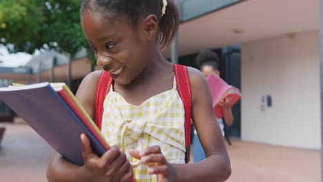Video-portrait-of-happy-african-american-schoolgirl-looking-at-her-books-outside-school