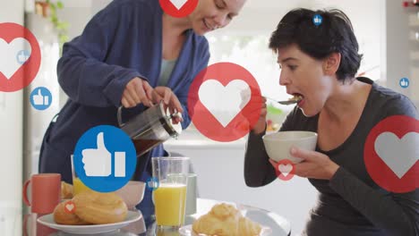Animation-of-hearts-and-like-icons-over-caucasian-female-couple-eating-breakfast
