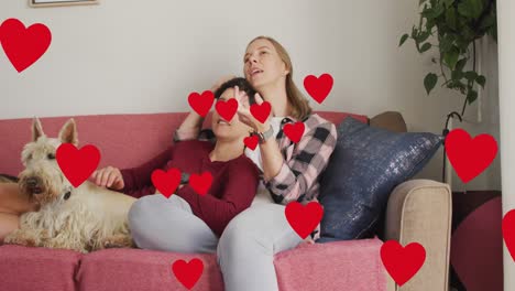 Animation-of-hearts-over-caucasian-female-couple-sitting-on-sofa-with-their-pet-dog