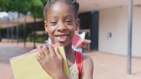Video-portrait-of-happy-african-american-schoolgirl-holding-her-books-outside-school