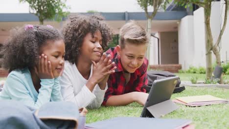 Video-of-three-happy-diverse-schoolchildren-watching-tablet-lying-in-schoolyard,-copy-space