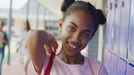 Video-portrait-of-happy-biracial-schoolgirl-smiling-by-lockers-in-school-corridor,-copy-space