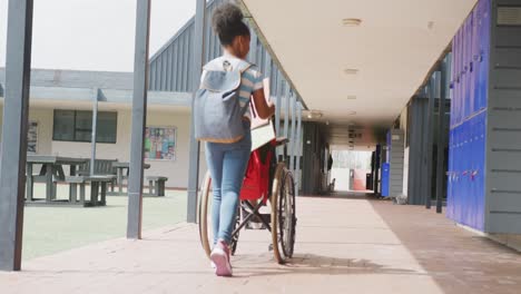 Video-rear-view-of-biracial-schoolgirl-pushing-wheelchair-in-school-corridor,-with-copy-space
