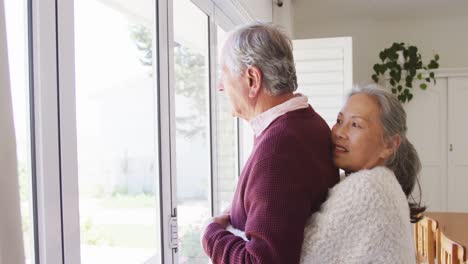 Glückliches,-Vielfältiges-älteres-Paar,-Das-Sich-Umarmt-Und-Gemeinsam-Durch-Das-Fenster-Schaut
