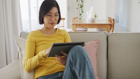 Glückliche-Asiatische-Frau,-Die-Ein-Tablet-Benutzt-Und-Auf-Der-Couch-Im-Wohnzimmer-Sitzt