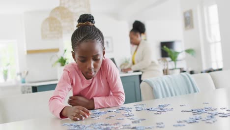Happy-african-american-granddaughter-doing-jigsaw-puzzle,-with-grandmother-in-background,-copy-space