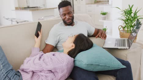 Diverse-couple-lying-on-couch-and-using-laptop-in-living-room