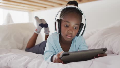 Happy-african-american-girl-in-headphones-using-tablet-lying-on-bed-at-home,-copy-space