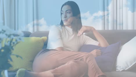 Animation-of-sky-with-clouds-over-biracial-woman-sitting-on-sofa