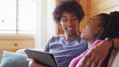 Happy-african-american-couple-using-tablet,-sitting-on-sofa-in-log-cabin,-slow-motion