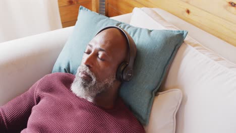 Happy-senior-african-american-man-in-log-cabin,-laying-on-sofa-and-using-headphones,-slow-motion
