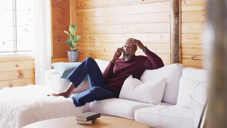 Happy-senior-african-american-man-spending-time-in-log-cabin,-sitting-on-sofa-and-using-smartphone