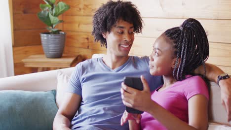 Happy-african-american-couple-using-smartphone,-sitting-on-sofa-in-log-cabin,-slow-motion