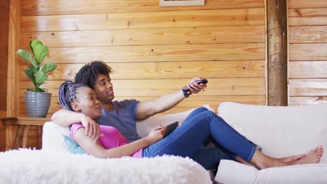 Happy-african-american-couple-watching-tv,-sitting-on-sofa-in-log-cabin,-slow-motion