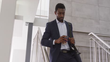 Happy-african-american-businessman-going-down-stairs-and-drinking-coffee-at-office,-slow-motion