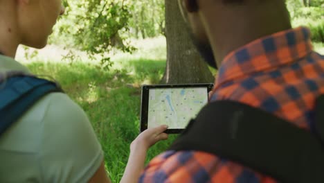 Diverse-couple-hiking-with-backpacks-and-using-tablet-with-map-in-park,-slow-motion