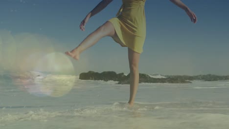 Animation-of-lens-flares-over-biracial-young-woman-with-arms-outstretched-walking-at-shore