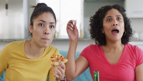 Two-diverse-happy-female-friends-having-beer-and-pizza-watching-tv-at-home,-in-slow-motion