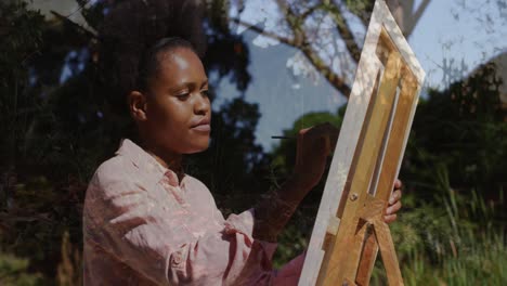 Animation-of-trees-over-african-american-woman-painting-on-easel-in-garden