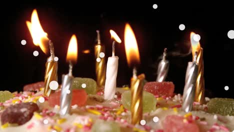 Animation-of-white-spots-floating-over-burning-candles-on-a-cake-against-black-background
