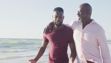 Video-of-african-american-senior-father-and-adult-son-walking-on-beach-and-talking