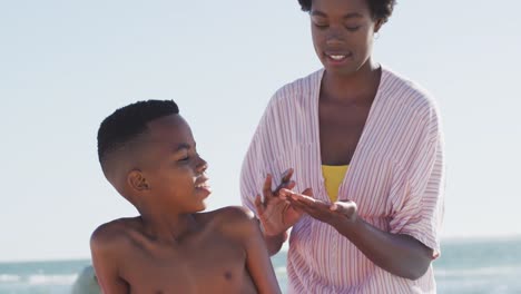 Video-Einer-Glücklichen-Afroamerikanischen-Mutter,-Die-Ihrem-Sohn-Am-Strand-Sonnencreme-Aufträgt