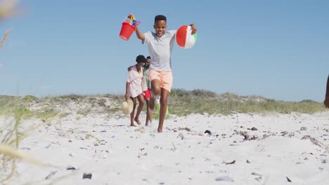 Glückliche-Afroamerikanische-Familie-Mit-Strandausrüstung,-Die-An-Einem-Sonnigen-Tag-Am-Strand-Spaziert
