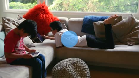 Animation-Eines-Netzwerks-Digitaler-Symbole-über-Zwei-Asiatische-Jungen,-Die-Zu-Hause-Auf-Der-Couch-Ein-Digitales-Tablet-Nutzen