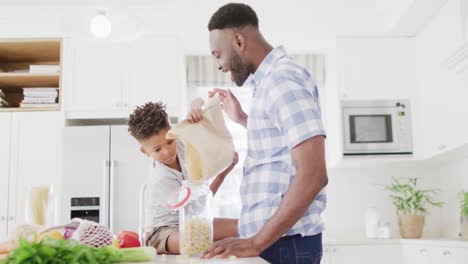 Feliz-Padre-E-Hijo-Afroamericanos-Llenando-Un-Frasco-De-Almacenamiento-Con-Pasta-En-La-Cocina,-Cámara-Lenta