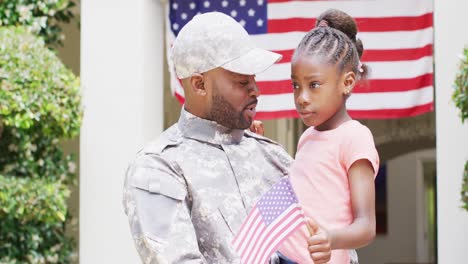 Happy-african-american-male-soldier-smiling-and-holding-daughter-with-flag-outside-home,-slow-motion
