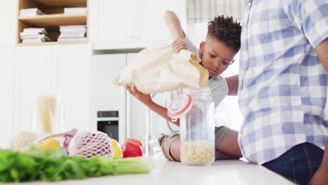 Padre-E-Hijo-Afroamericanos-Llenando-Tarro-De-Almacenamiento-Con-Pasta-En-La-Cocina,-Cámara-Lenta