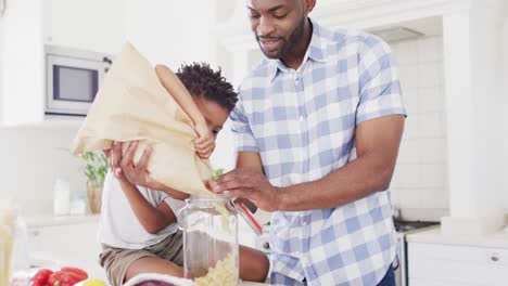 Feliz-Padre-E-Hijo-Afroamericanos-Llenando-Un-Frasco-De-Almacenamiento-Con-Pasta-En-La-Cocina,-Cámara-Lenta