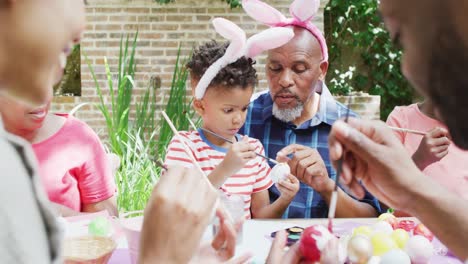 African-american-grandfather-and-grandson-painting-easter-eggs-with-family-in-garden,-slow-motion